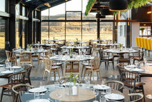 Load image into Gallery viewer, A large, modern restaurant interior with multiple round dining tables set with white plates and glassware, dark wood chairs, and black pendant lights. The space features a high glass ceiling, a bar area with bright yellow stools on the right, and a scenic outdoor landscape visible through the windows.